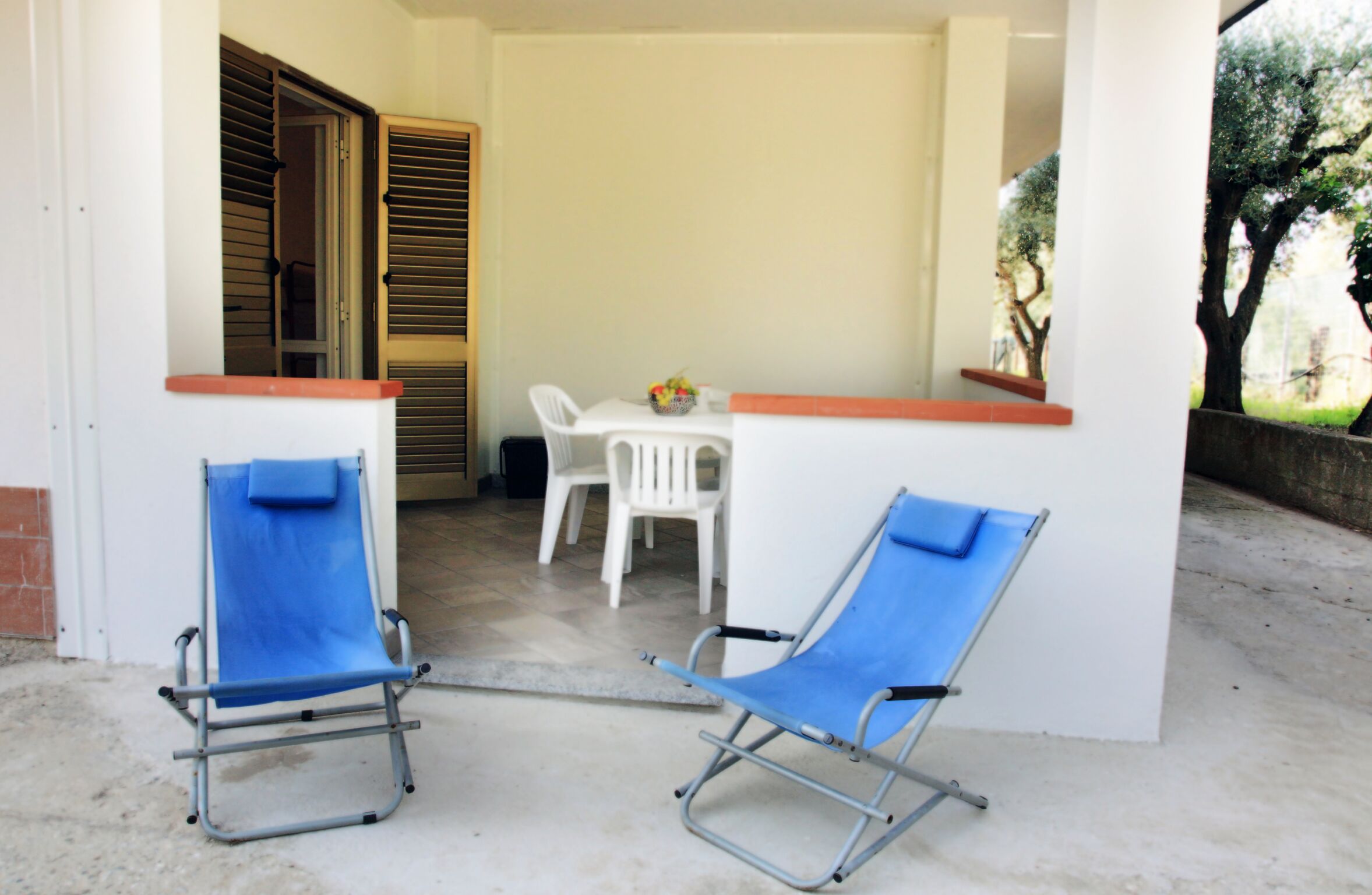 Un espace patio comprenant deux fauteuils bleus, une table blanche avec des chaises et une petite table agrémentée d’un arrangement floral.