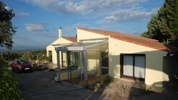 Une maison moderne dotée d’un patio entièrement vitré, une voiture rouge garée devant, et une vue dégagée sur la campagne environnante.