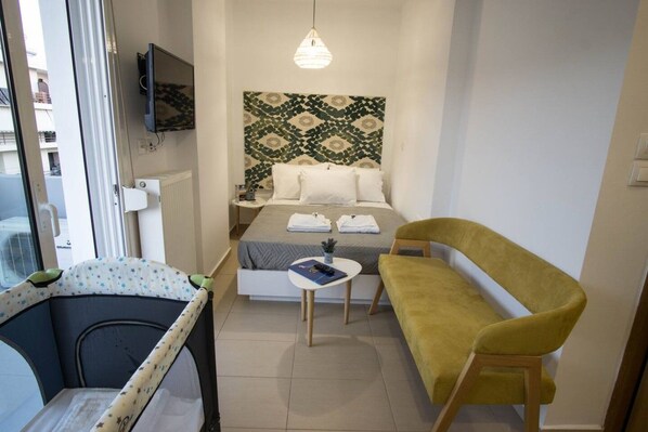 A compact hotel room with a bed, a small table, a yellow armchair, a television on the wall, and a patterned headboard.