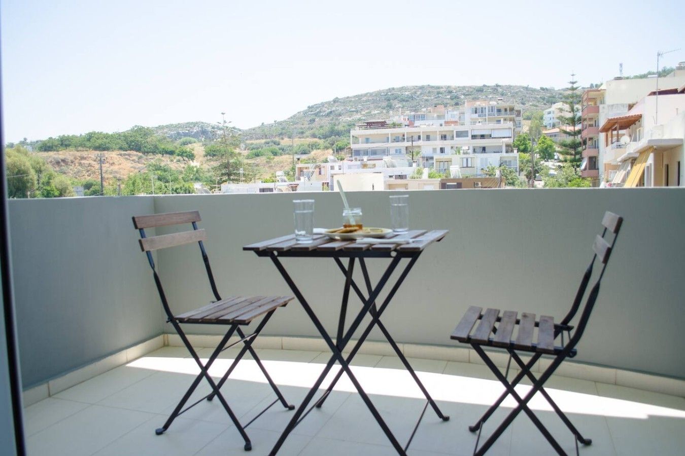 A balcony with a table and two chairs, overlooking a residential area with buildings and hills.
