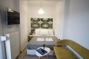 A compact hotel room with a bed, bedside table, a small table with a remote control, a yellow armchair, and a television mounted on the wall. - Urban Beach Sea (3C) (Rethymno)