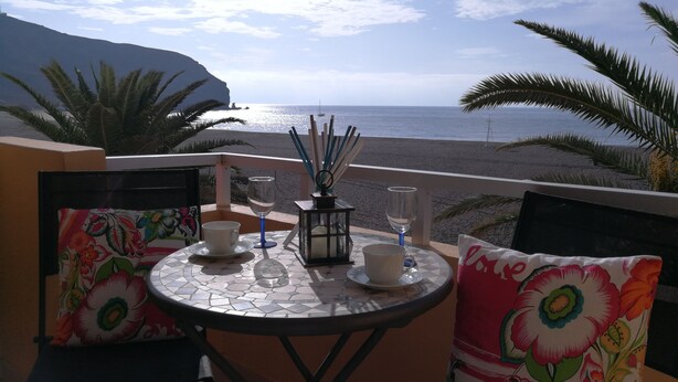 Un balcón con una mesa para dos, con vista a una playa y una montaña al fondo.