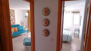 Un salón con un sofá azul, una mesa de centro blanca y un reposapiés azul. Se observan tres adornos en la pared y una ventana con cortinas.