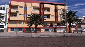 Un edificio de varios pisos con un letrero de clínica, palmeras y suelo arenoso al frente.
