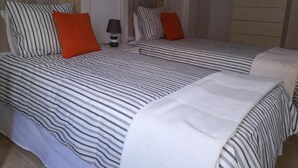 Una cama doble con ropa de cama a rayas y dos almohadas, una mesita de noche con lámpara y un aparador con un cuadro encima.