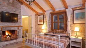 A bedroom with a stone fireplace, a bed with a metal frame, wooden beams, a television, and a window with curtains. - Quiet and Cozy Cottage in Nature (Buje)
