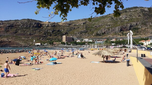 Een strand met mensen die zonnebaden en zwemmen, een rieten parasol en gebouwen op de achtergrond.