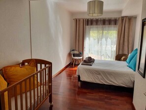 Une chambre avec un berceau en bois, un lit recouvert d’une couette blanche, une chaise et une vue sur l’extérieur par une fenêtre ornée de rideaux.