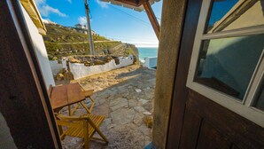 A stone patio with a wooden table and chair, a white wall, and a view of the sea and a hill with buildings. - Azenhas Beach Room - Azehas do Mar (Lisboa)
