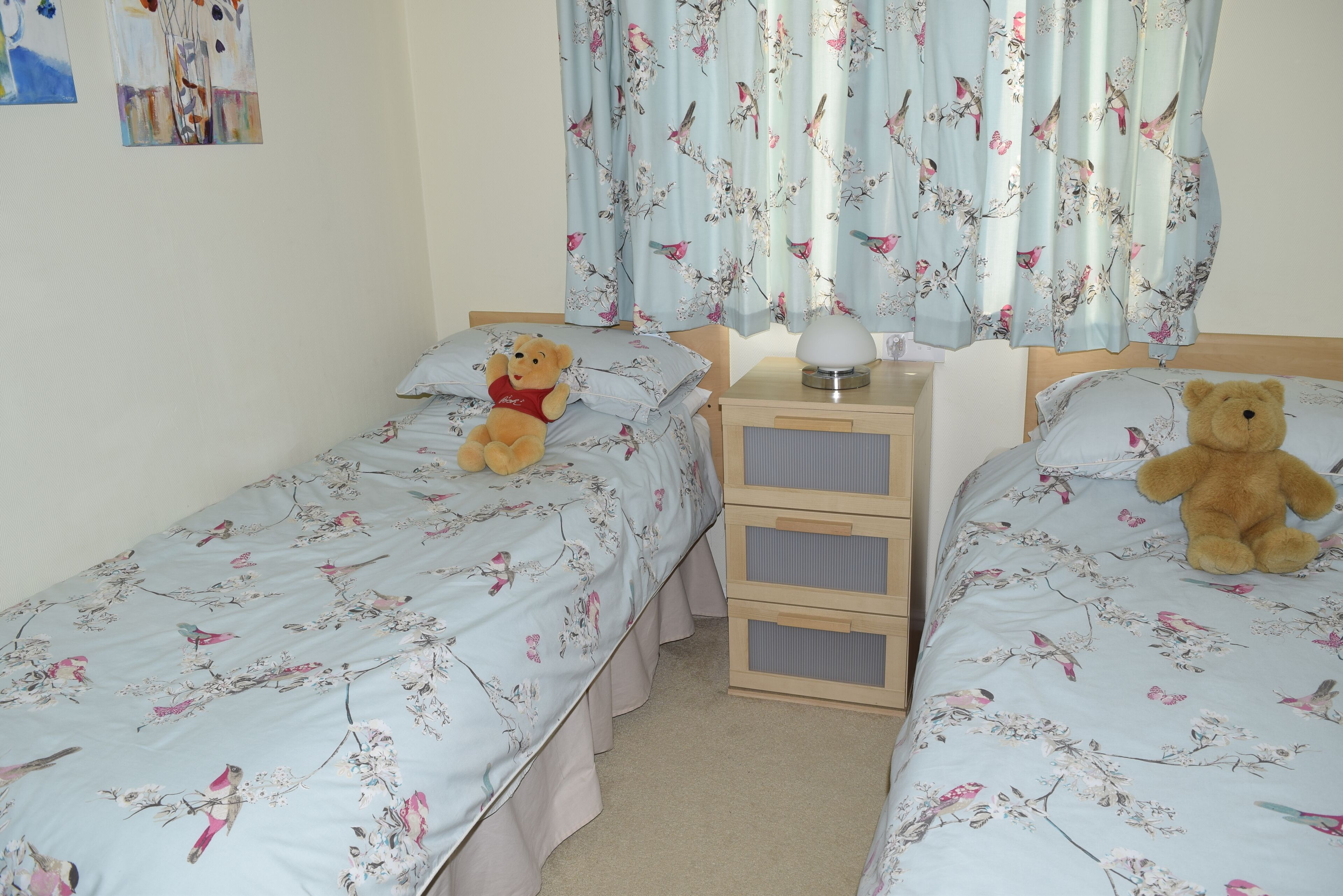 Une chambre avec deux lits, chacun agrémenté d’une literie à motifs floraux et d’un ours en peluche. Il y a deux tables de chevet avec des tiroirs et une lampe sur l’une d’elles.