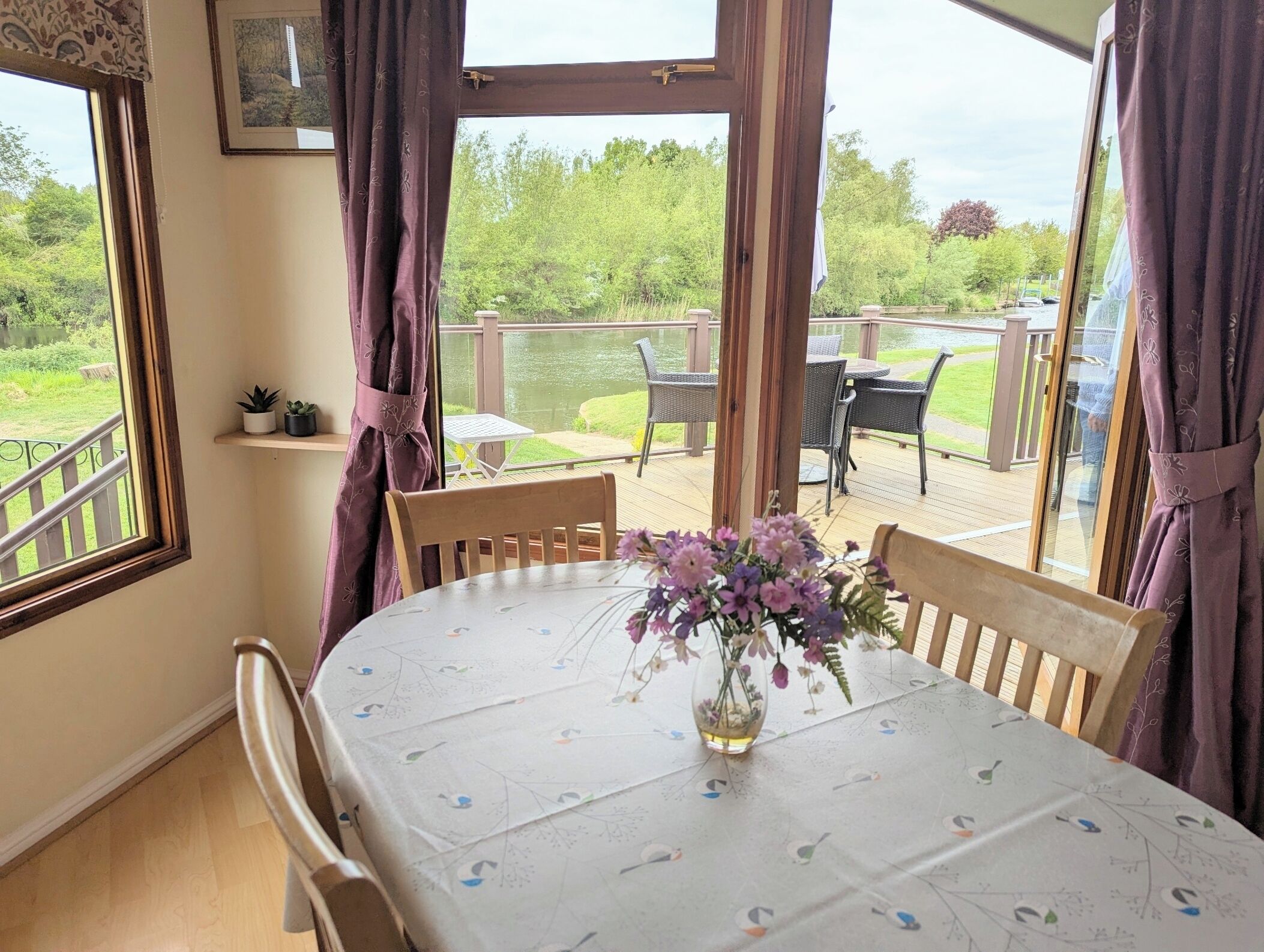 Een eethoek met een tafel gedekt voor twee personen, een vaas met bloemen en uitzicht op een terras en tuin door grote ramen.