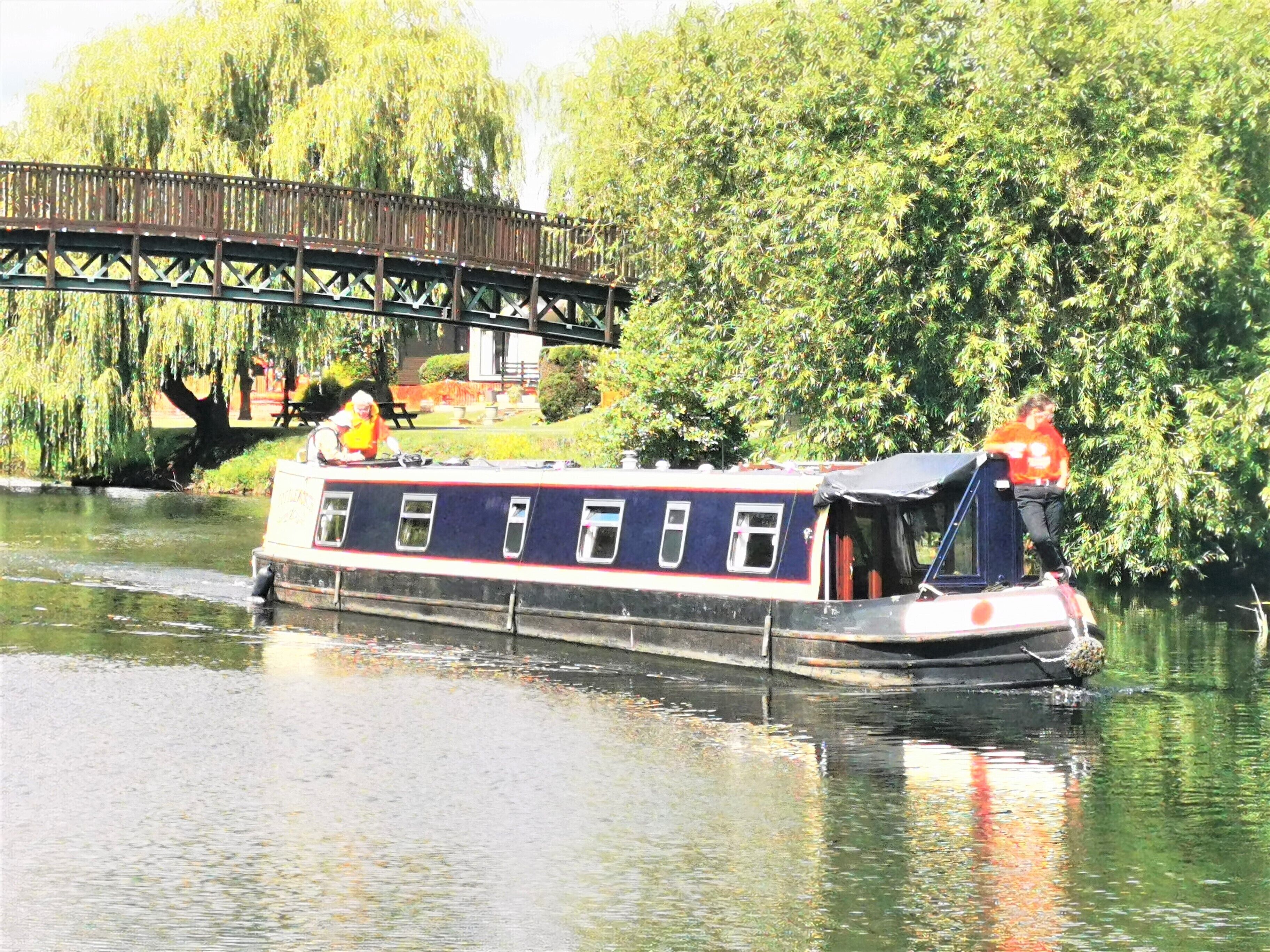 A narrowboat on a calm river with a bridge in the background.