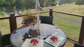 Une table avec un verre de vin, un bol de framboises et une plante en pot sur un balcon donnant sur une rivière.