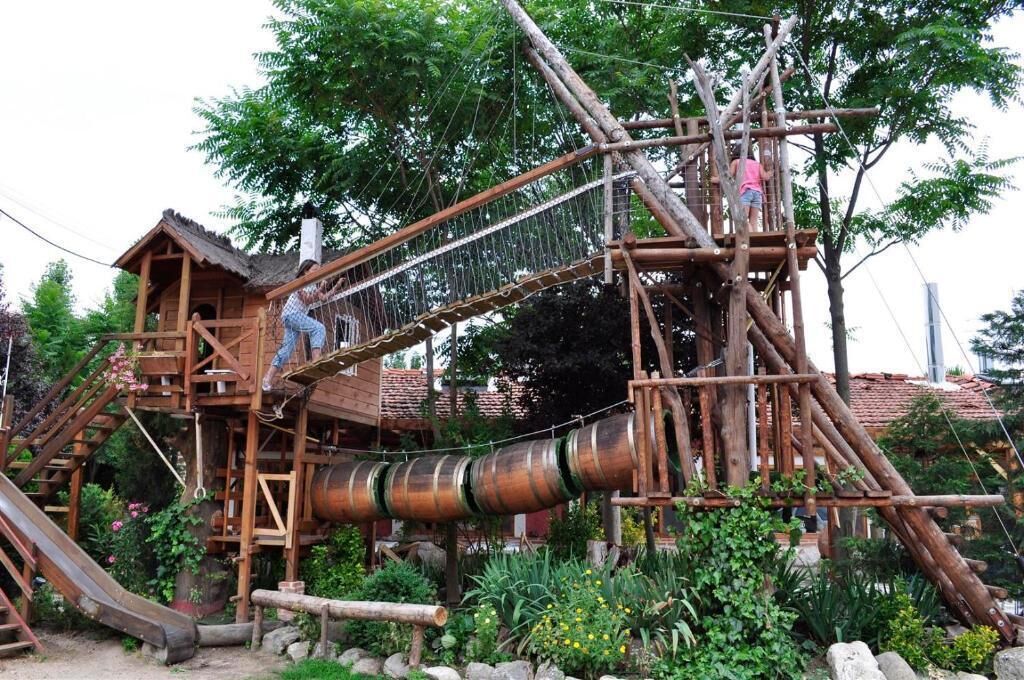 A wooden play structure with slides and climbing elements, surrounded by greenery.