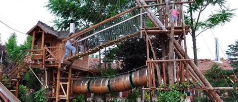 A wooden play structure with slides and climbing elements, surrounded by greenery.