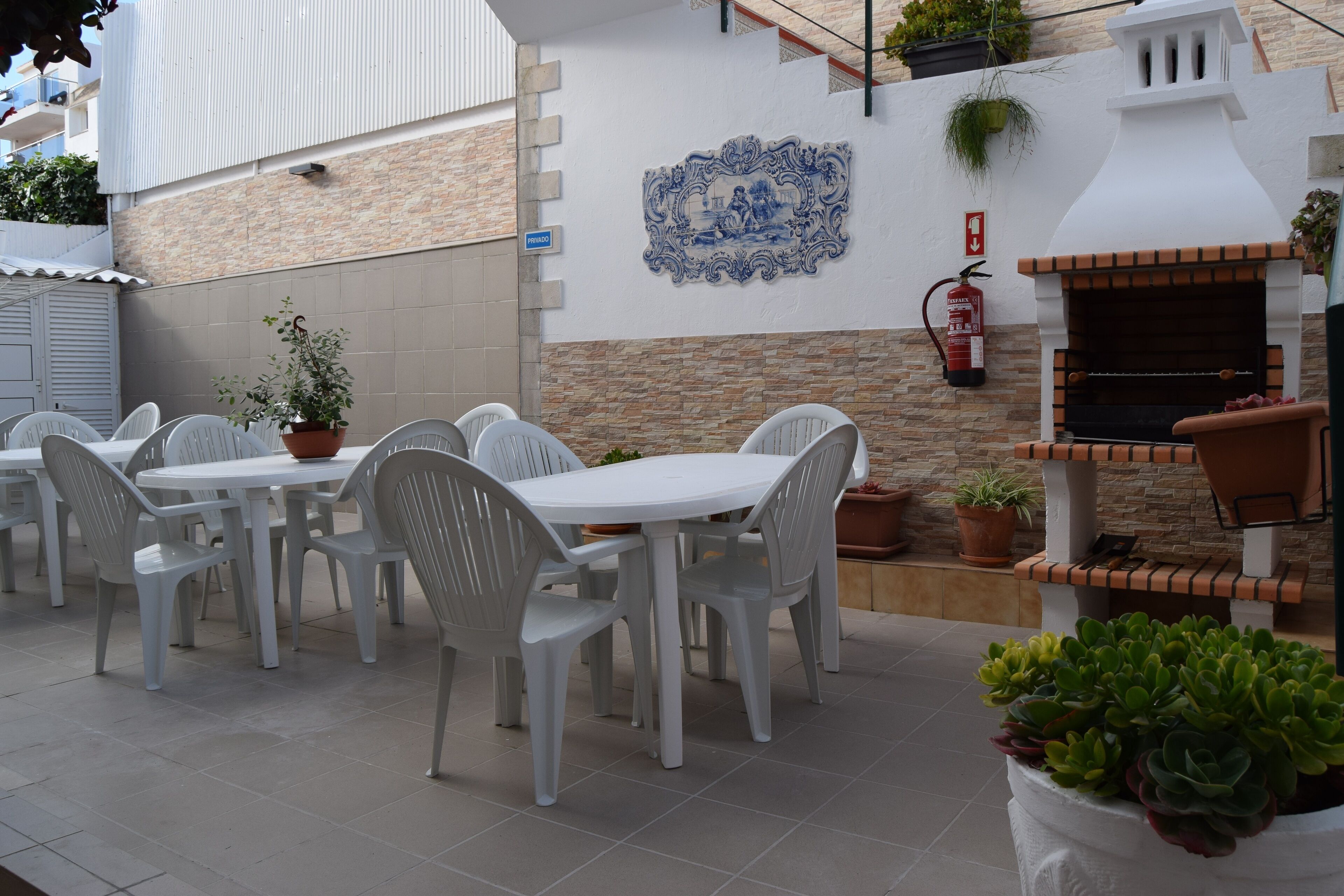 Een terras buiten met witte plastic stoelen en tafels, een bakoven en potplanten.
