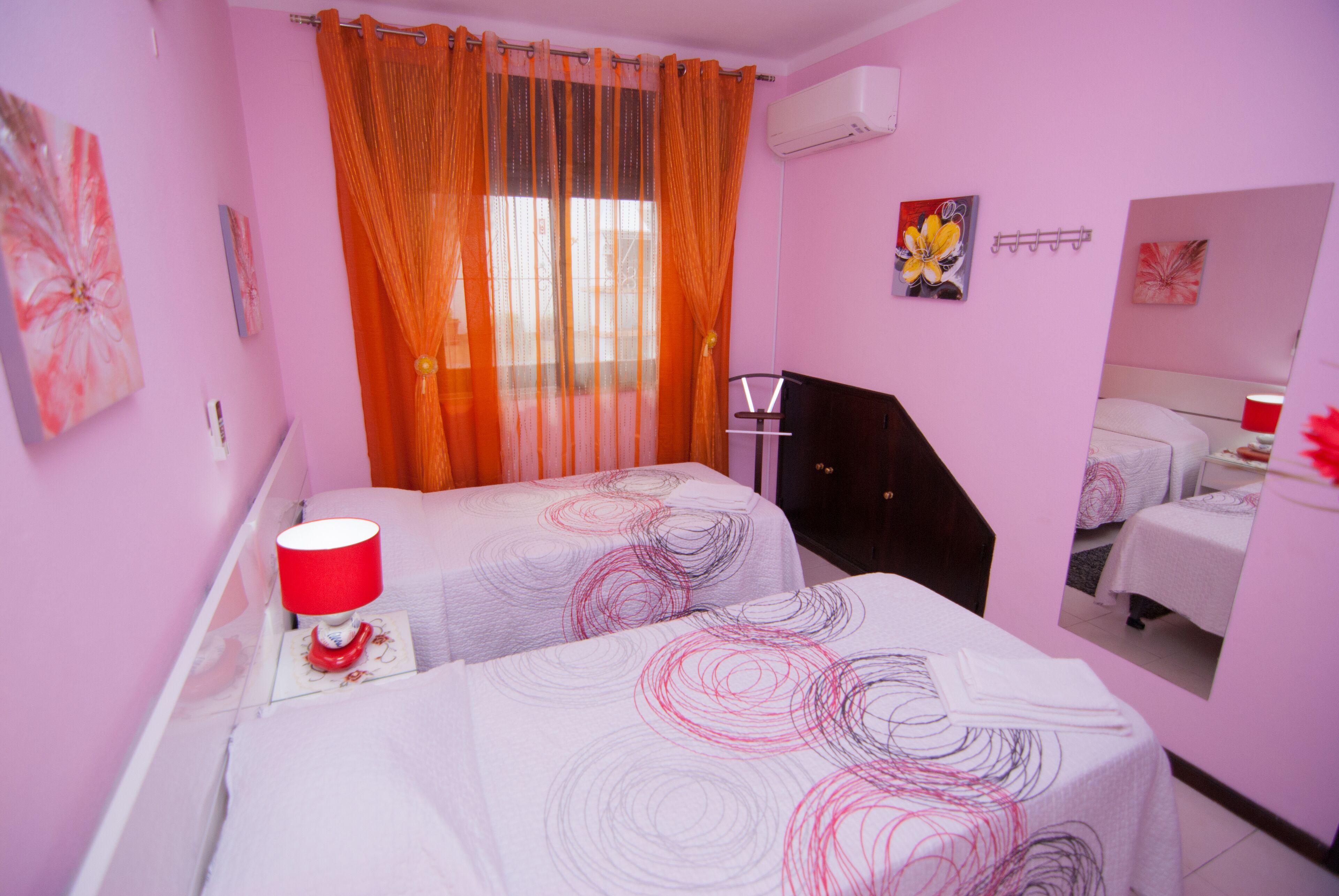 Habitación de hotel en tonos rosados, con dos camas, una mesita de noche con lámpara y obras de arte en las paredes.