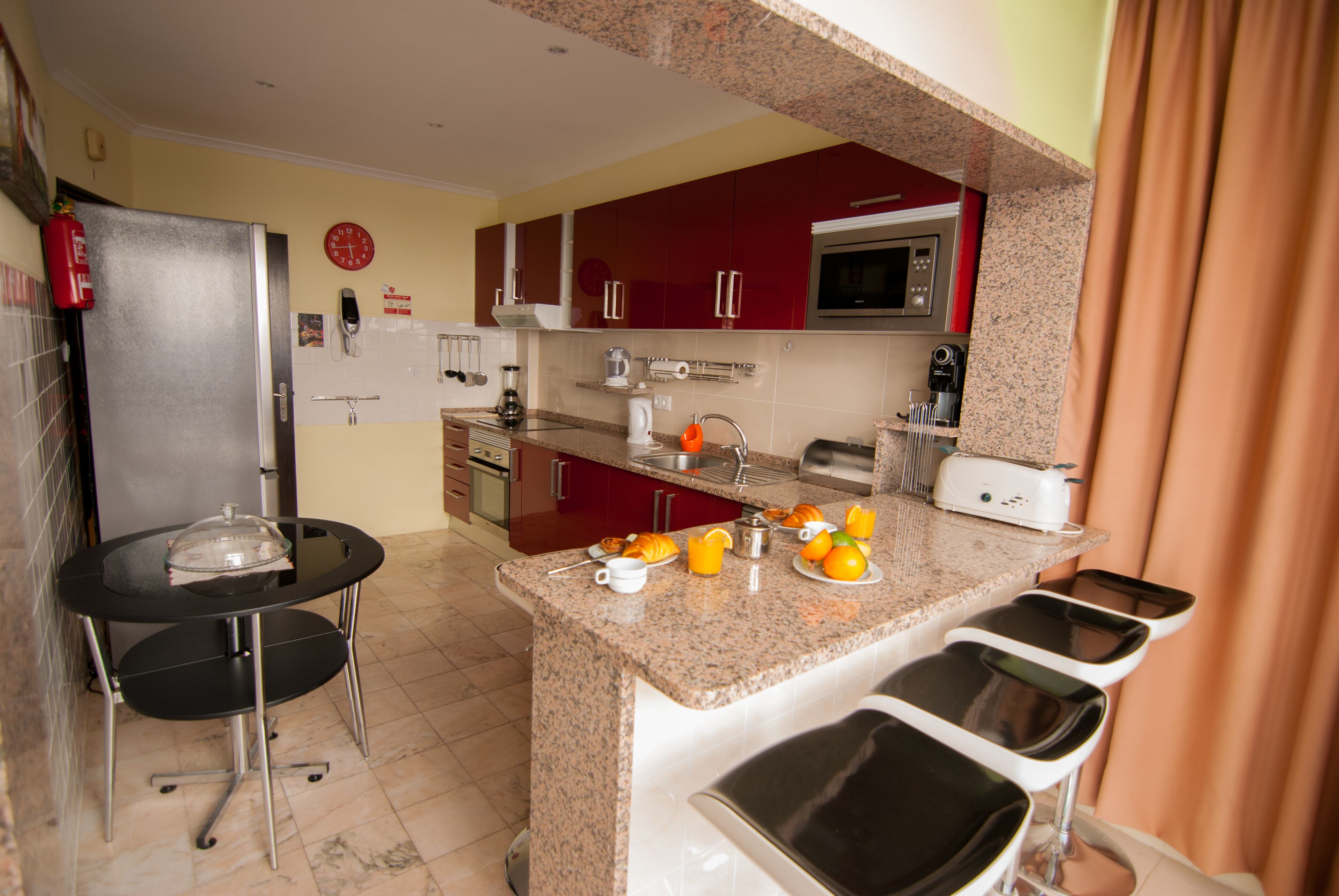 A compact kitchen with a refrigerator, microwave, and a coffee maker. There are three bar stools and a small table with a glass dome.