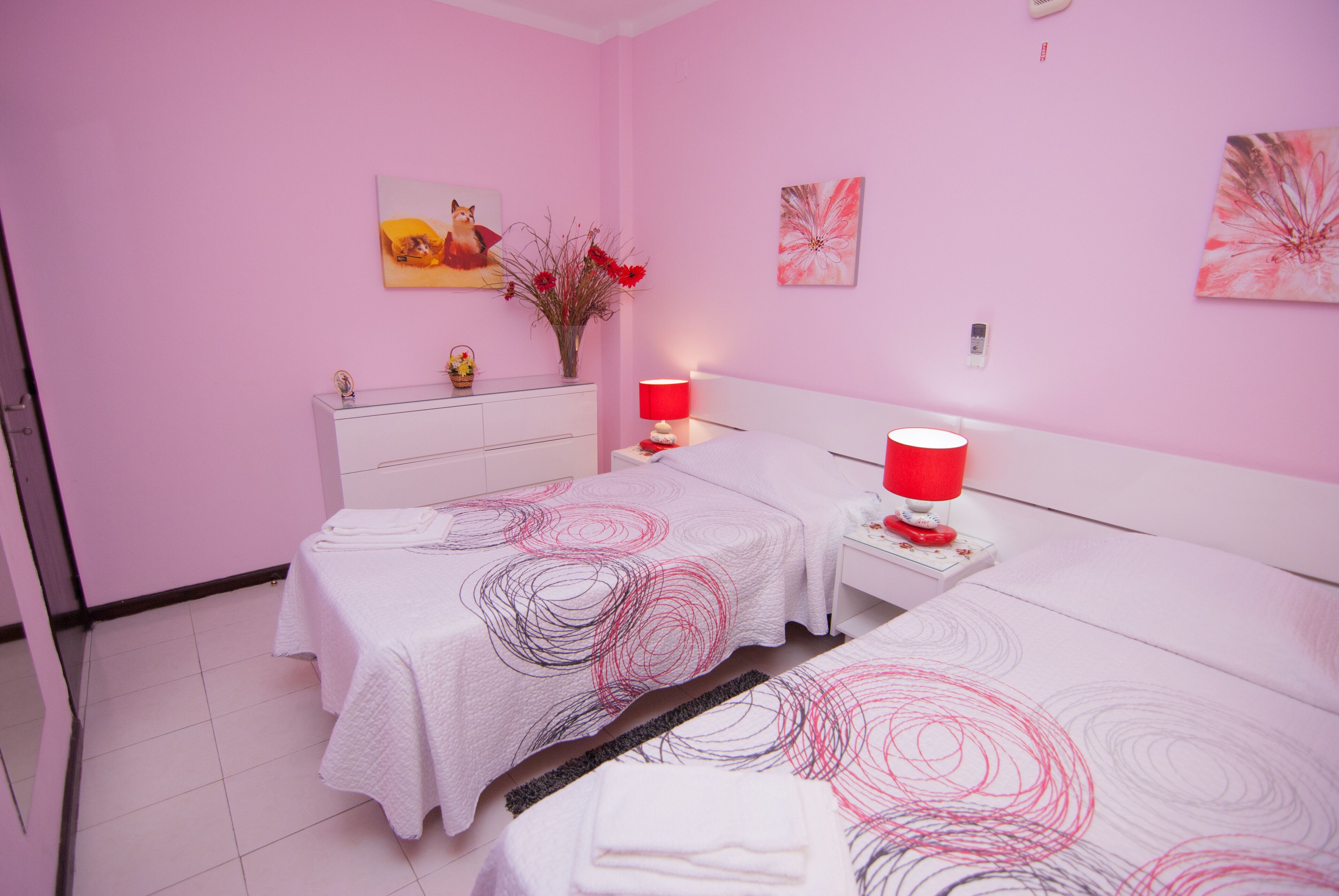 A pink-themed hotel room with two beds, a nightstand, a vanity, and artwork on the walls.