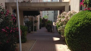 Un chemin pavé mène à l’entrée d’un bâtiment, de part et d’autre duquel s’étendent des haies soigneusement taillées et des plantes en fleurs.