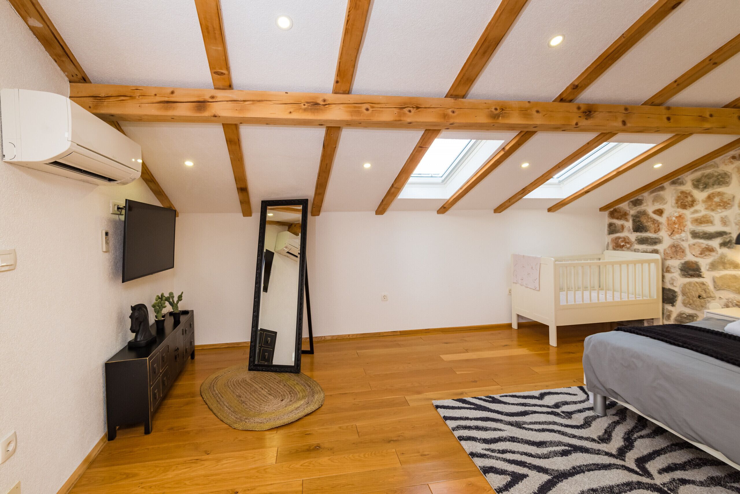 Une chambre avec un plafond en pente, des poutres en bois, un berceau, un lit, un miroir, une commode et un téléviseur.