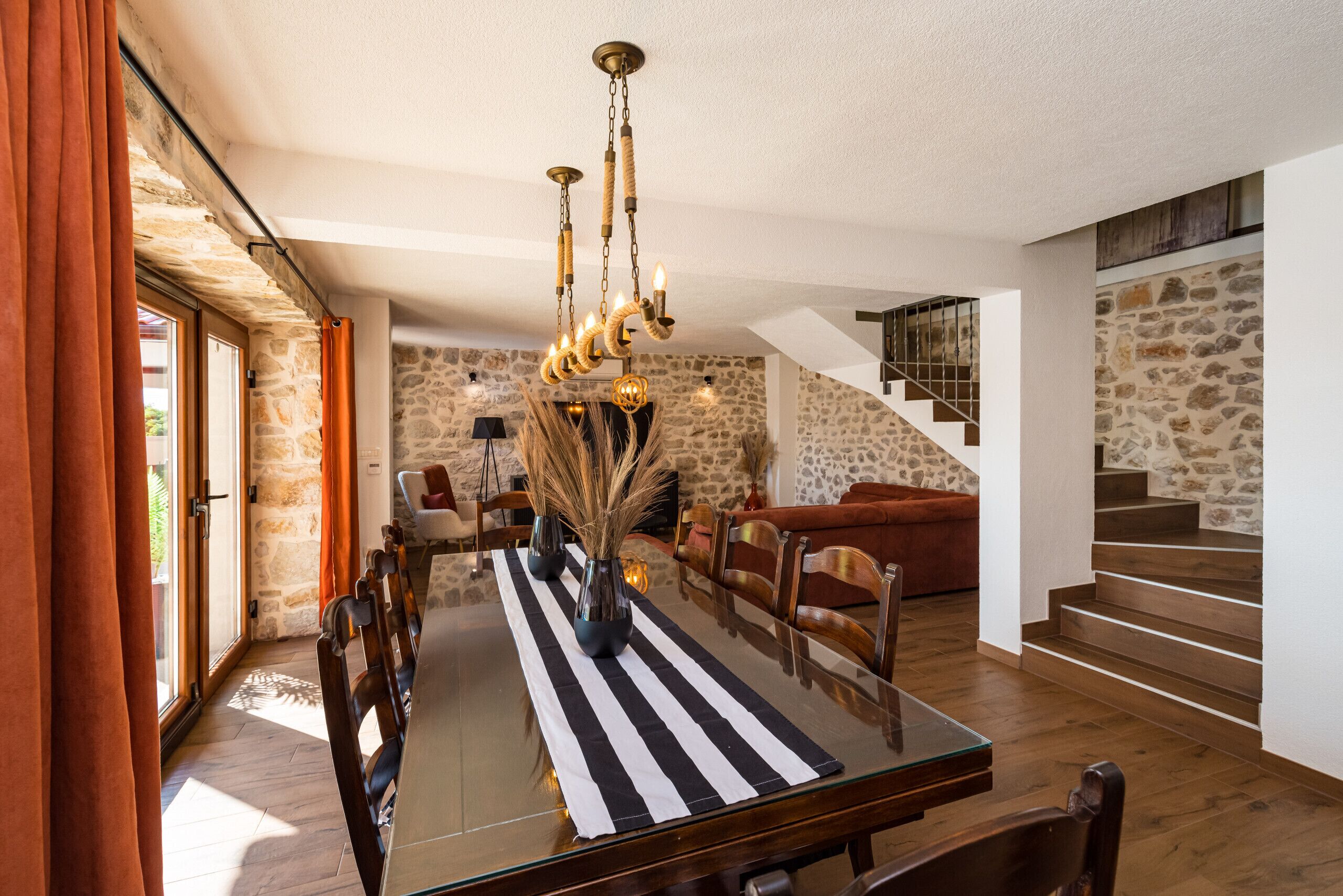 A dining area with a glass table, striped tablecloth, and a chandelier. There is a staircase with a stone wall in the background.