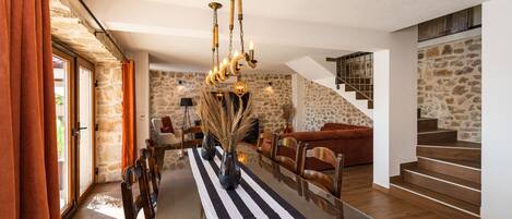 A dining area with a glass table, striped tablecloth, and a chandelier. There is a staircase with a stone wall in the background.