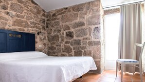 Een slaapkamer met een stenen muur, een bed met een blauw hoofdeinde, een stoel en een raam met gordijnen.