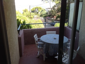 A small balcony with a round table and two chairs, overlooking a residential area with trees and a parked car. - Charming studio with sea view (Hyères)