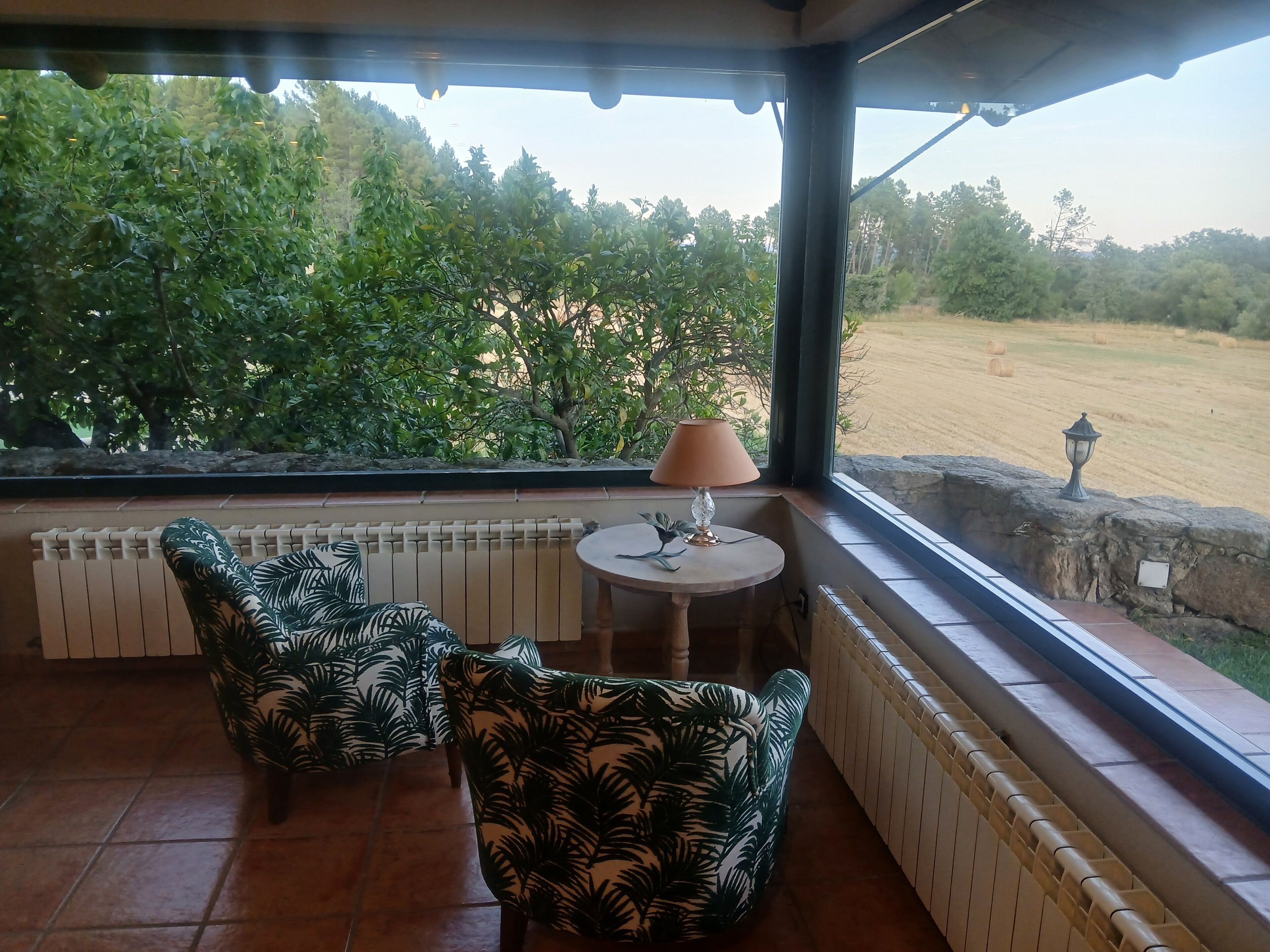 Eine überdachte Terrasse mit zwei Stühlen, einem Tisch und einer Lampe, mit Blick auf ein Feld mit Bäumen.