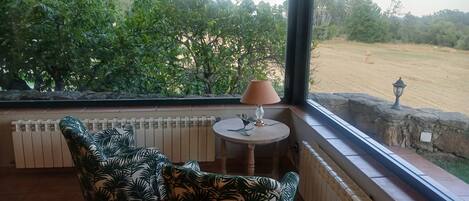Un porche cubierto con dos sillas, una mesa con una lámpara y vistas a un campo con árboles.