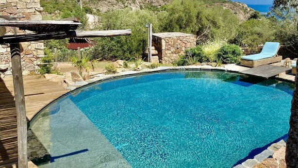 Une piscine d’un bleu éclatant, entourée de murs de pierre et d’une terrasse en bois, offrant une vue sur des collines et un ciel dégagé.