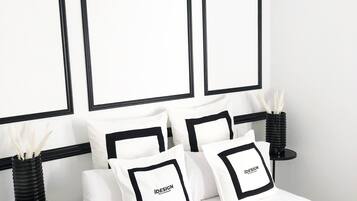 Une chambre à coucher avec un lit, du linge de lit blanc, des oreillers noirs ornés du mot « iDESIGN », des tables de chevet noires et une lampe noire.