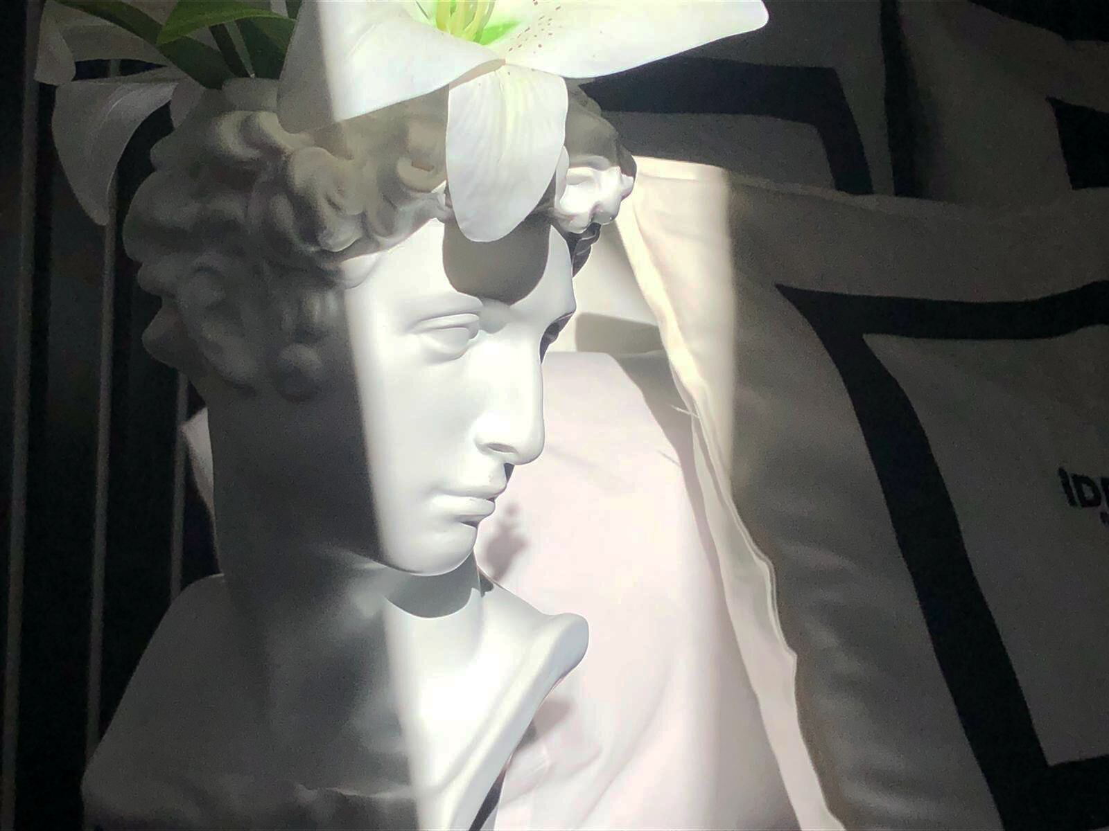 Eine weiße Büstenstatue mit einer Blume im Haar, vor dunklem Hintergrund mit einem Teilansicht eines schwarz-weißen Stoffes.