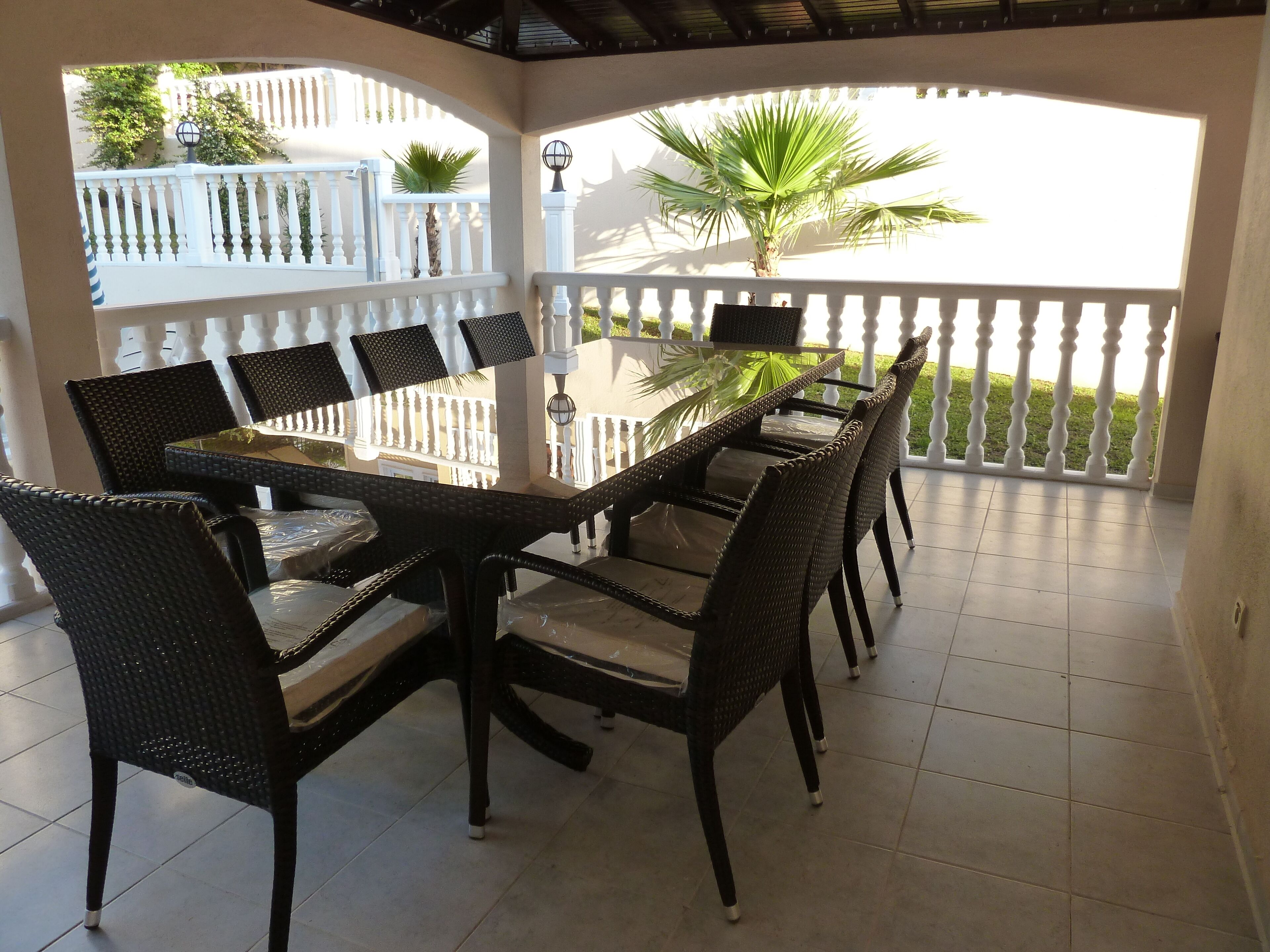 A covered outdoor dining area with a round table and wicker chairs, surrounded by a white balustrade and a view of a green lawn and palm tree.