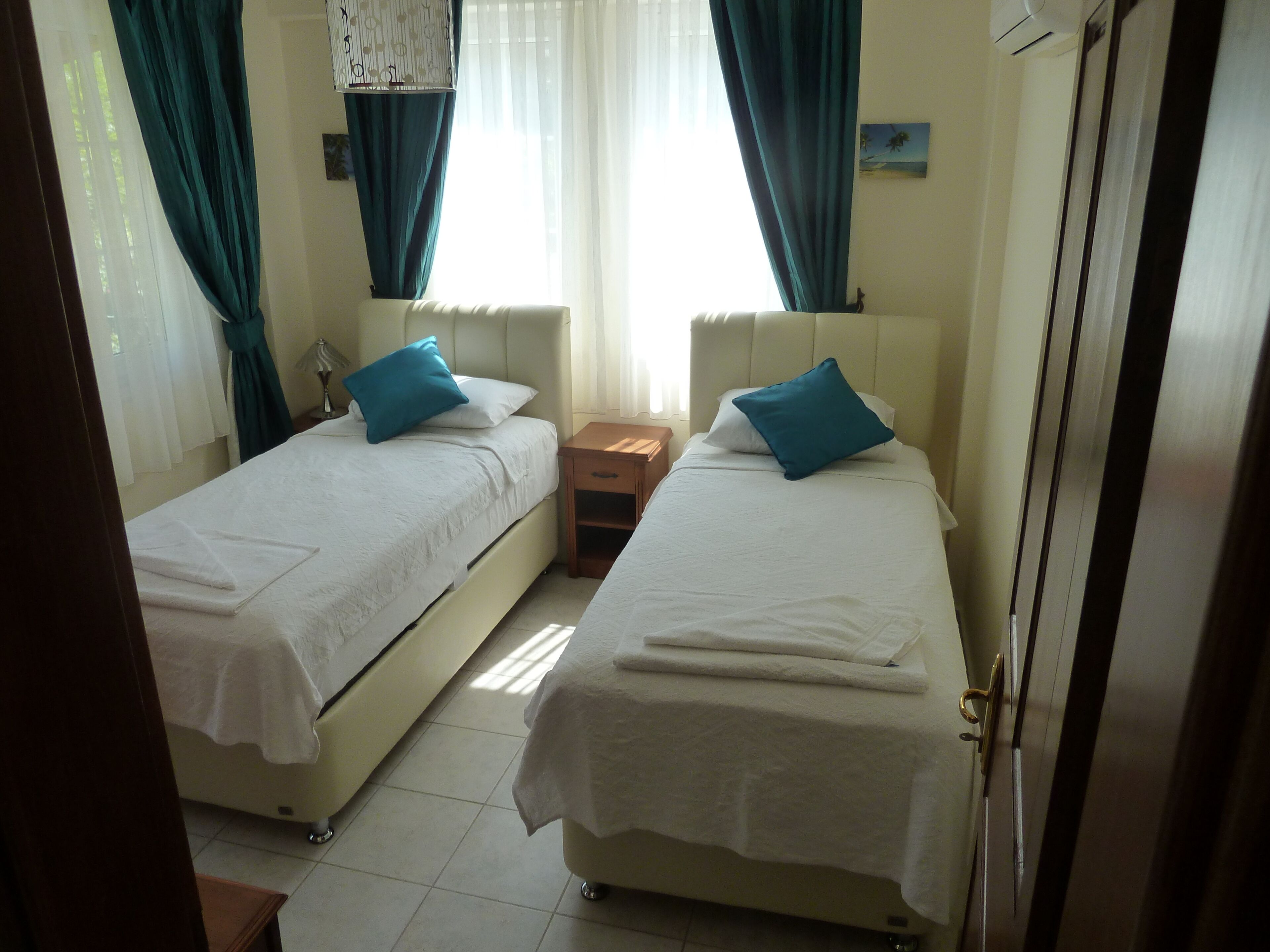 A room with two beds, each with a white bedspread and blue pillows, a wooden nightstand, and a window with curtains.