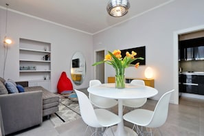A modern living room with a grey sofa, white chairs, a round table with a vase of flowers, and a kitchen area visible through an open door.