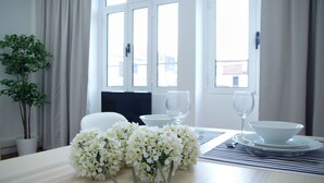 A dining area with a table set for two, featuring white plates, bowls, and wine glasses. A vase with white flowers is placed on the table. - Ribeira Funchal Center (Funchal)