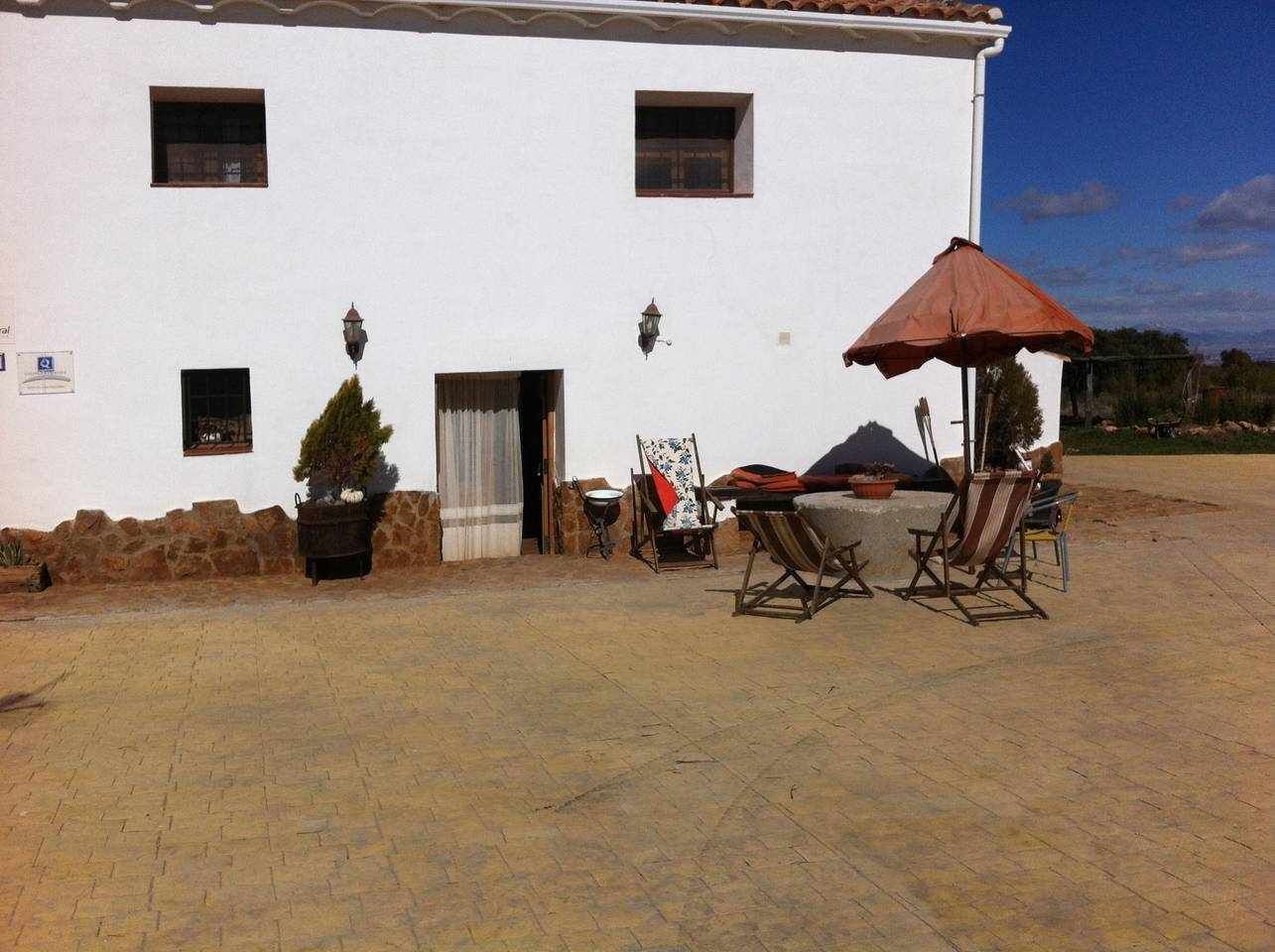 A white building with a paved patio, outdoor seating with a red umbrella, and a potted plant.