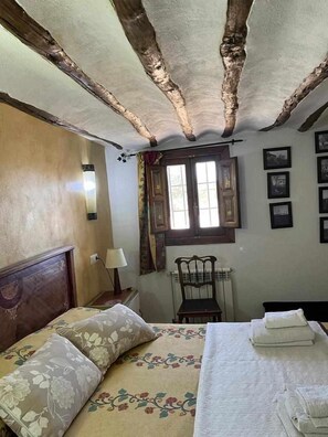 A bedroom with a wooden ceiling, a bed with floral bedding, a chair, and a window with curtains. - Cortijo rural bacares 1 (Freila)