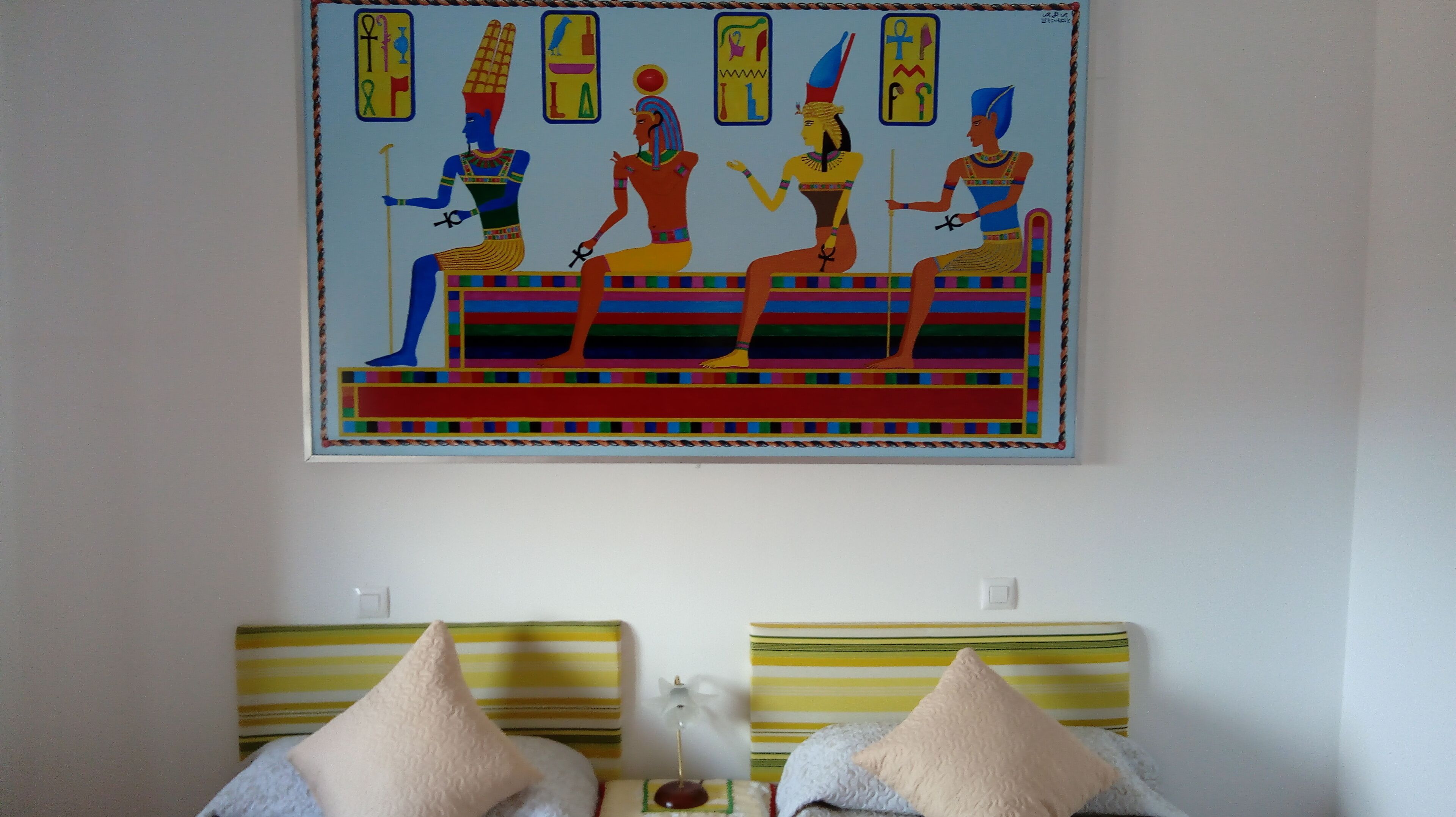 Un mur orné d’une peinture colorée représentant des figures égyptiennes et un lit avec des coussins à rayures.
