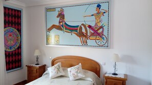 Un dormitorio con una cama, dos mesitas de noche, un cuadro colgado en la pared que representa una escena egipcia y una cortina estampada.
