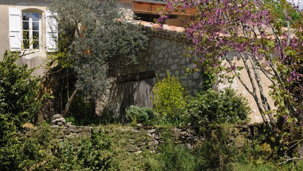 Una rustica casa in pietra con tetto in tegole, una porta di legno e un giardino con diverse piante e un albero in fiore.