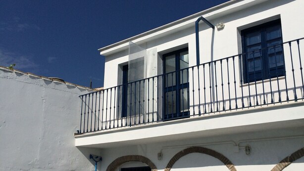 A two-story building with a white facade, a blue door, and a balcony with black railings. - Commander's corner (Tocina)