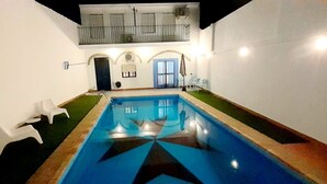 An indoor swimming pool with a geometric tile design, a small seating area with white plastic chairs, and a balcony with railings. - Commander's corner (Tocina)