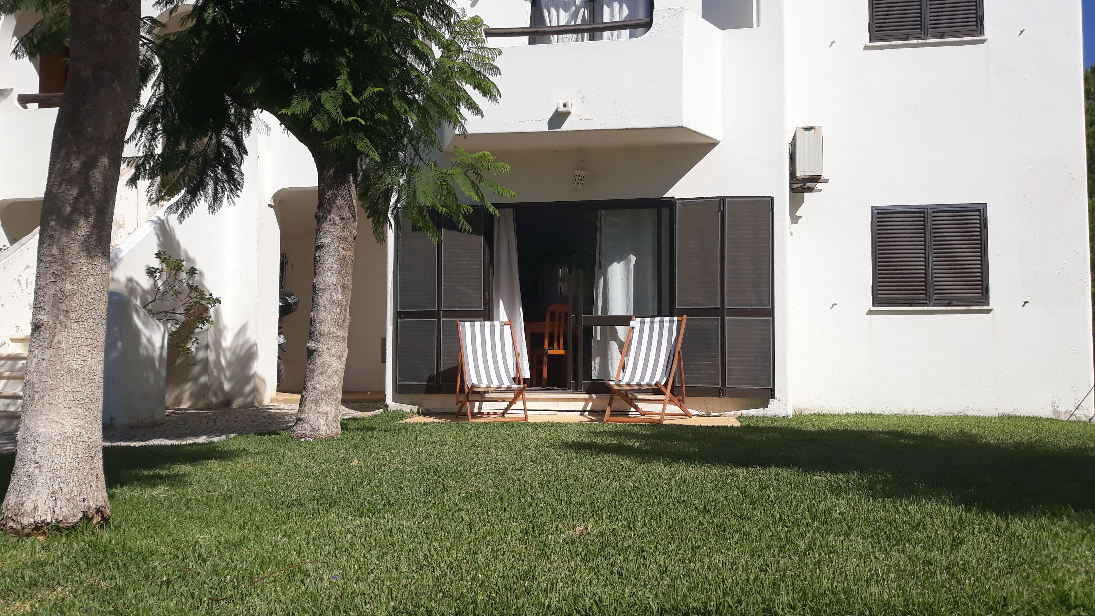 A white building with a balcony, two striped lounge chairs, and a tree in the foreground.