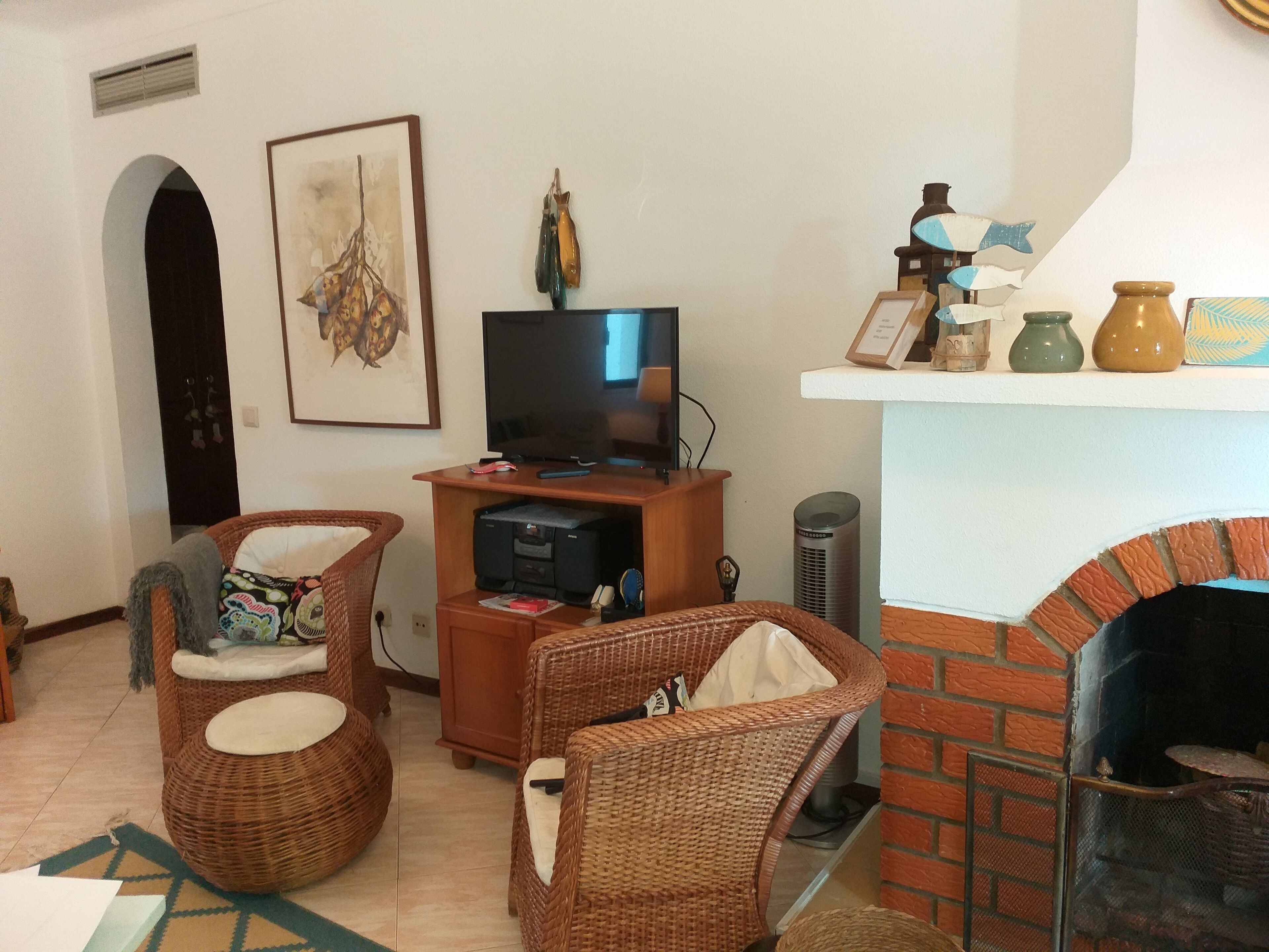Ein Wohnzimmer mit Korbmöbeln, einem Fernseher, einem Kamin und dekorativen Gegenständen auf Regalen.