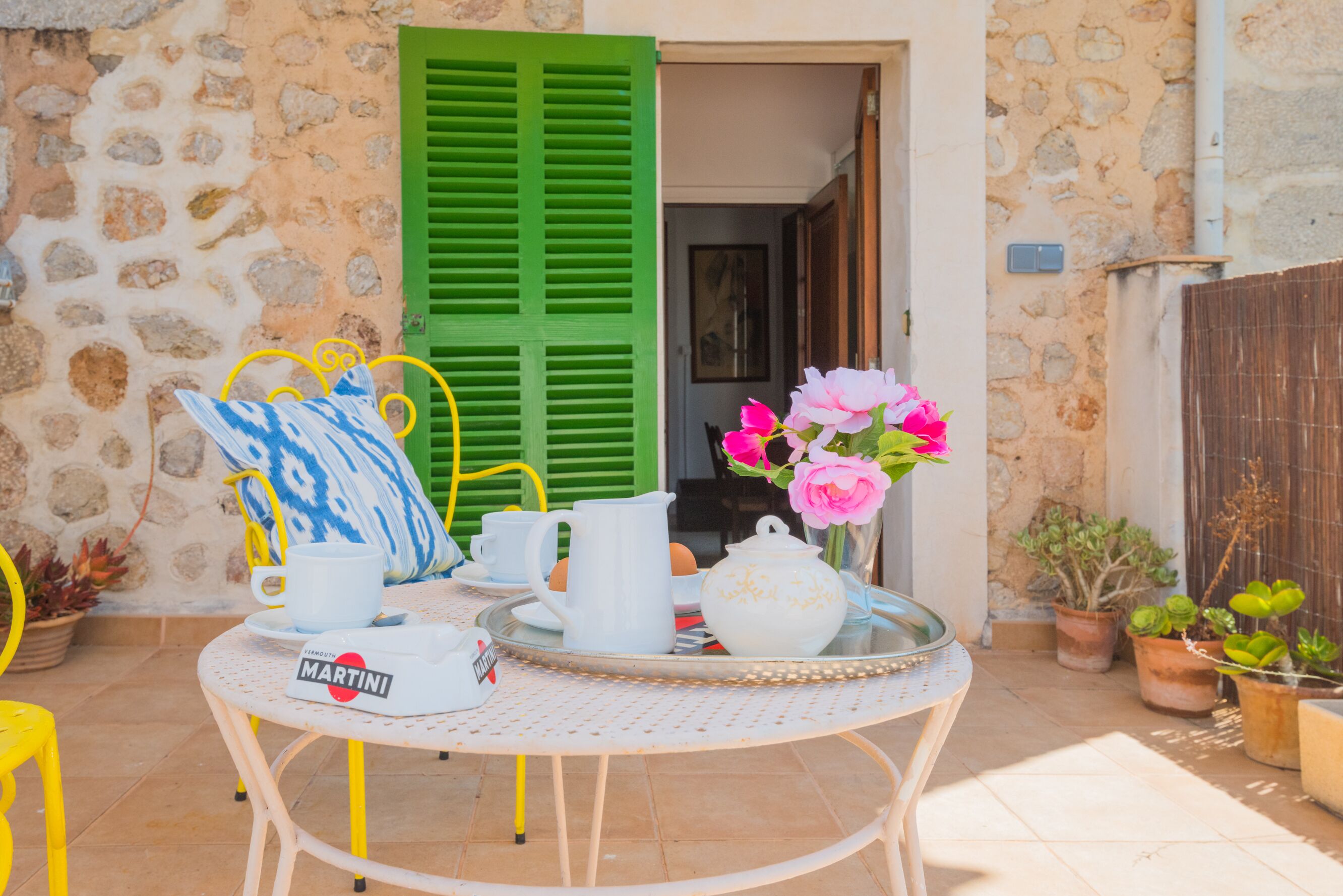 Une table ronde avec un plateau, une théière blanche, un pichet blanc et une tasse blanche accompagnée d’un verre à martini. On aperçoit, en arrière-plan, des plantes en pot et une porte à volets verts.