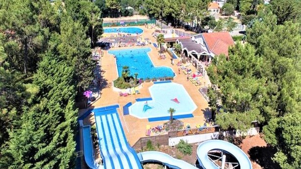 An aerial view of a resort with multiple swimming pools, a water slide, and a beach area surrounded by trees.