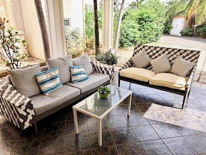A patio area with a sofa and armchair, a glass coffee table, and a view of a garden with plants and a building in the background.